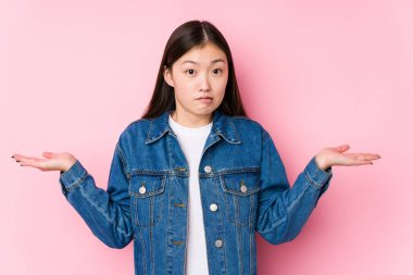 Pembe arka planda poz veren Çinli genç bir kadın. Kafası karışmış ve omuz silkiyor. Kopya alanını tutmak için..