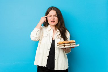 Genç, kıvrımlı bir kadın elinde kapkek tutuyor ve işaret parmağıyla hayal kırıklığı jesti yapıyor..