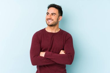 Mavi arka planda izole edilmiş genç beyaz adam kollarını kavuşturarak gülümsüyor..