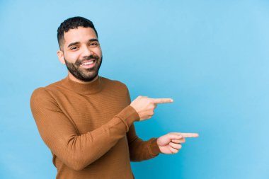 Mavi arka planda genç bir latin adam izole edilmiş. İşaret parmaklarıyla işaret ederek heyecanlı..