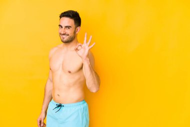 Sarı arka planda mayosu olan genç beyaz bir adam. Göz açıp kapayıncaya kadar Kafkasyalı genç bir adam. Elleriyle iyi bir jest yapıyor..
