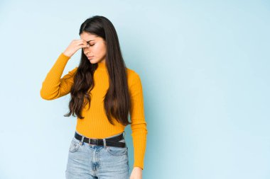 Genç Hintli bir kadın mavi arka planda izole edilmiş, başı ağrıyor, yüzüne dokunuyor..