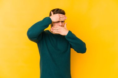 Sarı arka planda izole edilmiş genç beyaz adam kameraya parmaklarıyla göz kırpıyor, yüzünü kapatmaktan utanıyor..
