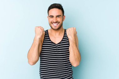 Kafkasyalı genç adam mavi arka planda izole edilmiş kaygısız ve heyecanlı tezahüratlar yapıyor. Zafer konsepti.