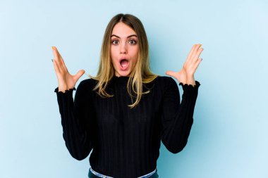 Mavi arka planda izole genç beyaz kadın zaferi ya da başarıyı kutluyor, şaşırıyor ve şok oluyor..