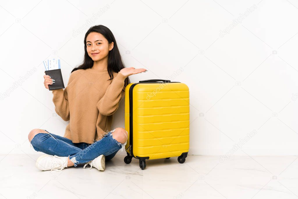 Young chinese traveler woman sittting on the floor with a suitcase isolated showing a copy space on a palm and holding another hand on waist.