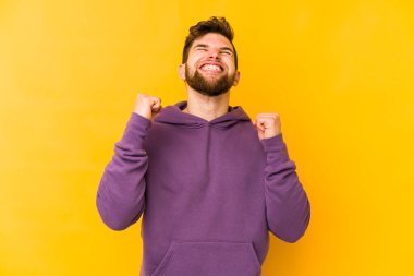 Sarı arka planda izole edilmiş genç beyaz adam zaferi, tutkuyu ve coşkuyu kutluyor, mutlu bir ifade..