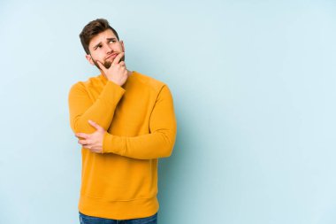 Kafkasyalı genç adam mavi arka planda izole edilmiş düşünüyor ve bakıyor, yansıtıyor, düşünüyor, bir fantezi kuruyor..