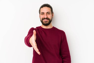 Genç, uzun saçlı bir adam beyaz bir arka planda izole edilmiş. Kameraya elini uzatarak selam veriyor..