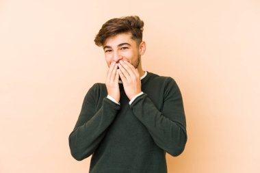 Genç Arap bir adam bej bir arka planda izole edilmiş. Bir şeye gülüyor, elleriyle ağzını kapatıyor..