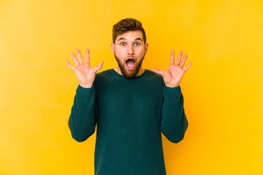 Sarı arka planda izole genç beyaz adam hoş bir sürpriz alıyor, heyecanlı ve el kaldırıyor..