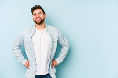 Kafkasyalı genç, mavi arka planda izole edilmiş. Kendine güvenen, kalçaları tutan biri..