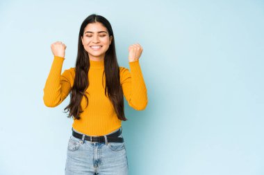 Genç Hintli kadın mavi arka planda izole edilmiş kaygısız ve heyecanlı tezahüratlar yapıyor. Zafer konsepti.