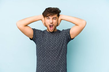 Genç Arap bir adam mavi bir arka planda izole edilmiş kulakları kulaklarıyla kapatıyor çok fazla ses duymamaya çalışıyor..