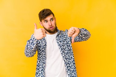 Sarı arka planda izole edilmiş genç beyaz adam baş parmağını kaldırıyor ve baş parmak aşağı, zor seçim konsepti.
