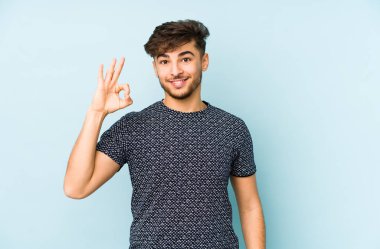 Genç Arap bir adam mavi bir arka planda izole edilmiş neşeli ve kendinden emin bir şekilde iyi bir jest yapıyor..