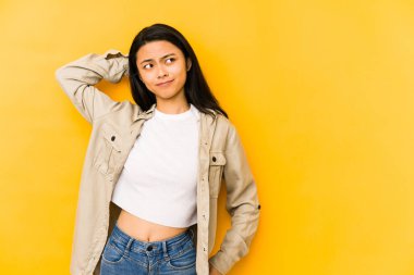 Sarı bir arka planda izole edilmiş genç Çinli kadın kafasının arkasına dokunuyor, düşünüyor ve bir seçim yapıyor..