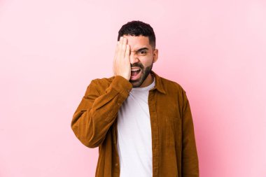 Pembe arka planda genç bir latin adam yalnız başına yüzün yarısını avuç içiyle kaplayarak eğleniyor..