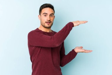 Mavi arka planda izole genç beyaz adam şok ve hayret içinde elleri arasında bir kopya boşluk tutuyor.