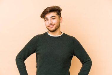 Genç Arap bir adam bej bir arka planda izole edilmiş. Kendine güvenen, kalçaları tutan..