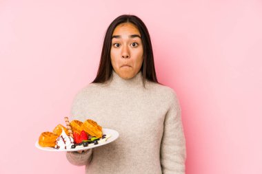 Genç Asyalı bir kadın waffle tutuyor, omuz silkiyor ve gözleri açık..