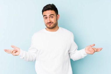 Kafkasyalı genç adam mavi arka planda izole edilmiş sorgulama jestinde şüphe ediyor ve omuz silkiyor..