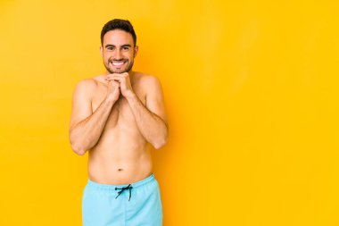 Sarı arka planda mayo giyen genç, beyaz tenli, elleri çenesinin altında olan genç bir adam, mutlu bir şekilde yan tarafa bakıyor..