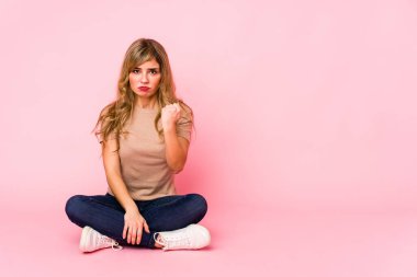 Pembe bir stüdyoda oturan genç sarışın beyaz kadın kameraya yumruğunu gösteriyor, agresif yüz ifadesi..