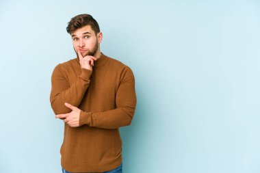 Mavi arka planda izole edilmiş genç beyaz adam şüpheli ve şüpheci bir ifadeyle yana bakıyor..