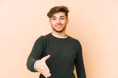 Bej bir arka planda izole edilmiş genç bir Arap adam selamlama jesti olarak kameraya elini uzatıyor..