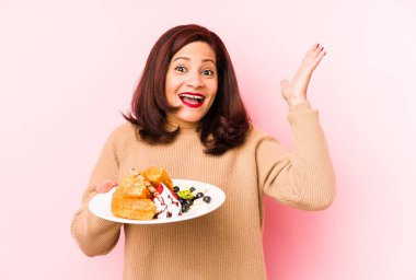Orta yaşlı, waffle tutan Latin bir kadın hoş bir sürprizle karşılaşıyor, heyecanlı ve el kaldırıyor..