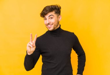 Sarı arka planda izole edilmiş genç bir Arap zafer işareti gösteriyor ve gülümsüyor..