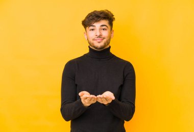 Sarı arka planda izole edilmiş genç bir Arap adam elinde avuç içi bir şeyle kameraya sunuyor..