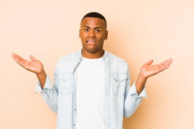 Bej arka planda izole edilmiş genç Latin bir adam. Kopya alanını tutmak için kafası karışık ve omuz silkiyor..