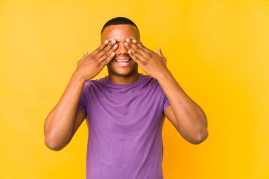 Sarı arka planda izole edilmiş genç latin adam elleriyle gözlerini kapatıyor, geniş bir gülümsemeyle bir sürpriz bekliyor..