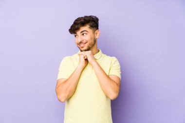 Mor arka planda izole edilmiş genç bir Arap adam ellerini çenesinin altında tutuyor ve mutlu bir şekilde yan tarafa bakıyor..