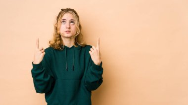 Beyaz tenli genç bir kadın, bej arka planda izole edilmiş, ağzı açık, üst tarafı işaret ediyor..