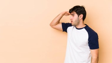 Genç adam bej arka planda izole edilmiş. Uzağa bakıyor. Elini alnına koyuyor..