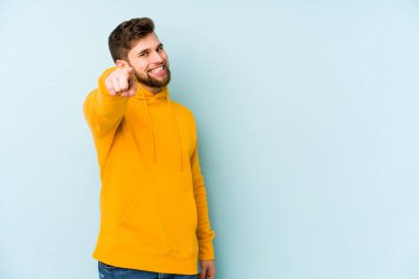 Mavi arka planda izole edilmiş genç beyaz adam parmaklarıyla ön tarafı işaret ediyor..