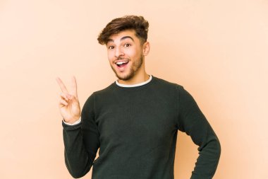 Genç Arap adam bej bir arka planda izole edilmiş neşeli ve kaygısız parmakları olan bir barış sembolü gösteriyor..