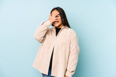 Mavi arka planda izole edilmiş genç bir kadın kameraya parmaklarıyla göz kırpıyor, yüzünü kapatmaktan utanıyor..