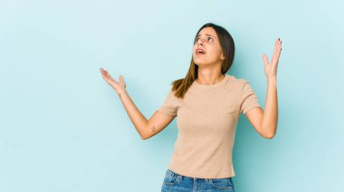 Mavi arka planda izole edilmiş genç bir kadın gökyüzüne haykırıyor, yukarı bakıyor, hayal kırıklığına uğramış..