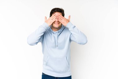 Beyaz arka planda soyutlanmış genç bir adam elleriyle gözlerini kapatıyor, geniş bir gülümsemeyle bir sürpriz bekliyor..