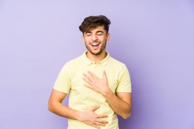 Mor arka planda izole edilmiş genç bir Arap adam mutlu bir şekilde gülüyor ve ellerini karnının üstünde tutarak eğleniyor..