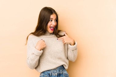 Beyaz tenli genç bir kadın bej arka planda izole edilmiş. Parmakla göstererek şaşırmış, geniş bir gülümsemeyle..