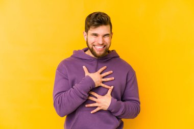 Sarı arka planda izole edilmiş genç beyaz adam mutlu bir şekilde gülüyor ve ellerini karnının üstünde tutarak eğleniyor..