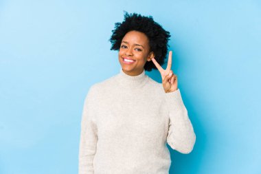 Orta yaşlı Afrikalı Amerikalı kadın mavi arka planda iki numarayı parmaklarıyla gösteriyor..