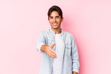 Pembe arka planda poz veren genç beyaz adam kameraya el uzatarak selam veriyor..