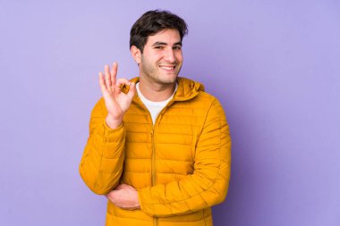 Mor arka planda izole olmuş genç bir adam bir göz kırpıyor ve elleriyle iyi bir jest yapıyor..