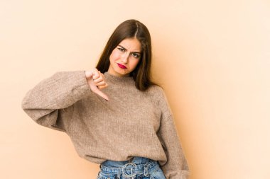 Beyazlı genç bir kadın bej arka planda izole edilmiş, hoşlanmadığı bir jest gösteriyor, başparmakları aşağıda. Anlaşmazlık konsepti.